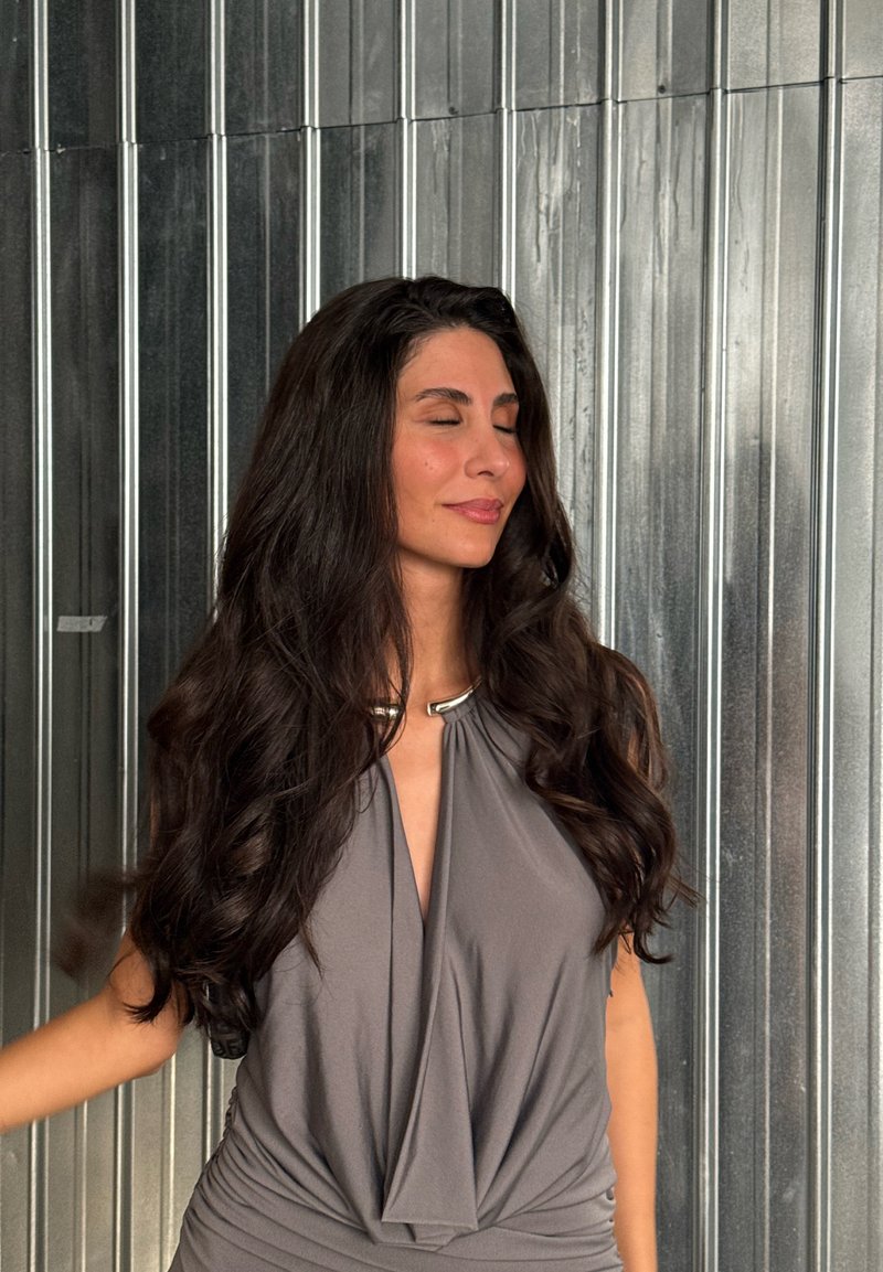 Haut gris drapé avec un accent métallique au col, présentant un tissu doux et fluide avec des détails froncés. Les cheveux sont longs et ondulés.