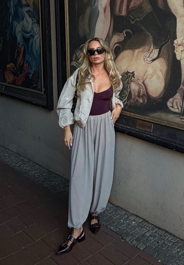Mujer con gafas de sol y pantalones beige holgados está de pie contra una pared con grandes pinturas clásicas.
