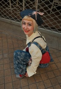 Joven con cabello rubio corto, vestido con un mono de denim, camisa crema, sombrero a rayas, bolso rojo y zapatos plateados, agachado en el pavimento de baldosas, sonriendo.