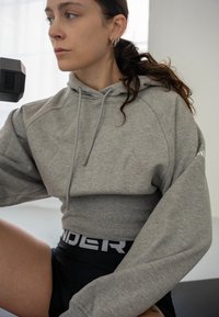 Jeune femme en sweat à capuche gris court et short noir soulevant un haltère, se préparant à faire de l'exercice dans un espace intérieur lumineux.