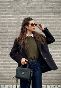 Crni oversized kaput preko maslinasto zelene veste, bijele košulje i tamnih traperica. Drži malu crnu torbicu. Nosive tamne sunčane naočale.