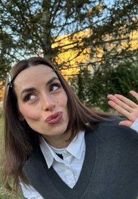 Jeune femme aux cheveux foncés et pinces à cheveux, faisant la moue et levant la main en plein air, avec des arbres et un bâtiment baigné de soleil en arrière-plan.