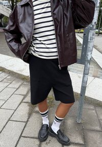 Donker bordeaux nepleer jas, gestreepte zwart-wit shirt, zwarte knie-lange rok, stevige zwarte loafers en sportieve witte sokken met zwarte strepen.