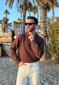 Homme portant des lunettes de soleil, un pull marron et un pantalon blanc, debout sur une plage rocheuse avec des palmiers et une clôture en bois en arrière-plan.