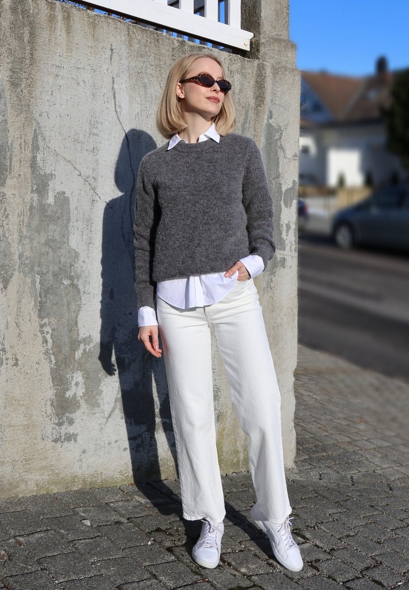 Femme aux cheveux blonds portant des lunettes de soleil, un pull gris par-dessus une chemise blanche, un pantalon blanc et des baskets blanches, debout contre un mur en béton.