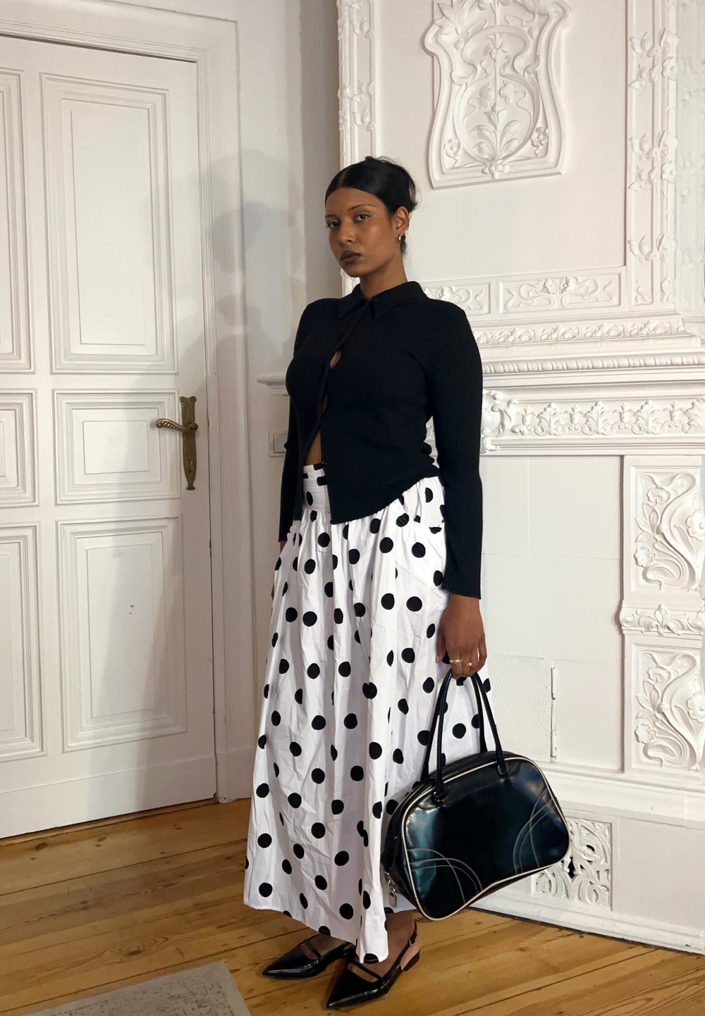 Cardigan preto com nervuras e decote abotoado, combinado com uma saia maxi branca com bolinhas pretas, sapatos de salto baixo pretos e uma mala preta.