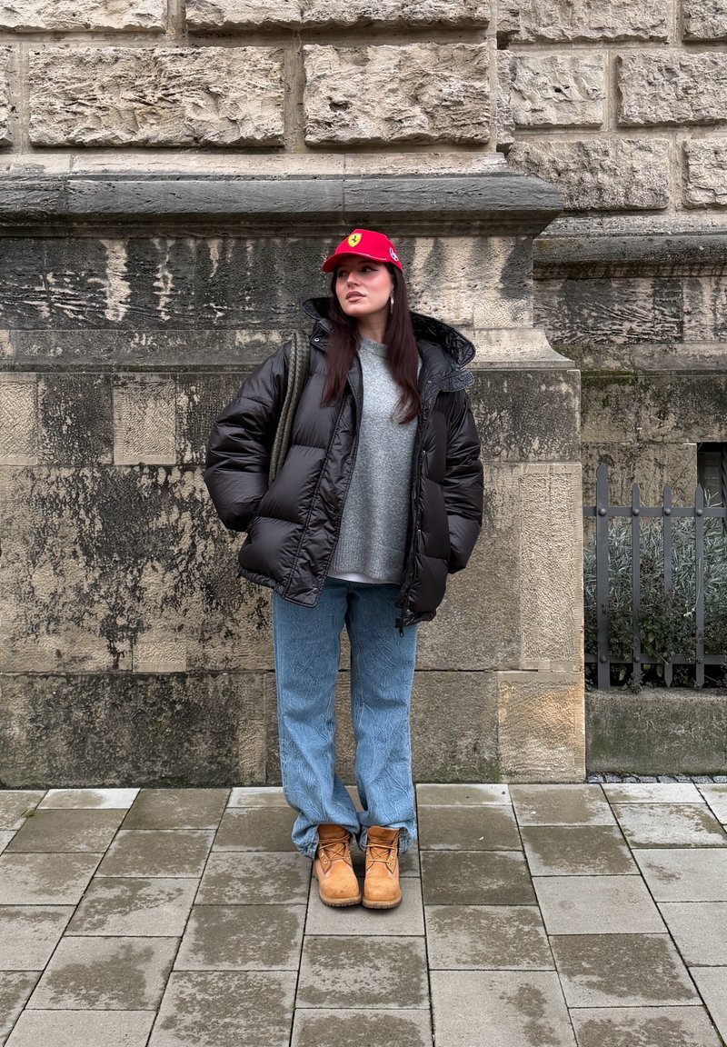 Doudoune noire, pull gris, jean bleu clair et bottes beige. Casquette rouge avec logo ; debout contre un mur de pierre sur des pavés.