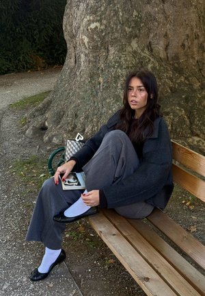 Pantalones de pierna ancha grises, suéter negro, calcetines blancos y bailarinas negras. Sosteniendo un libro, sentado en un banco de madera cerca de un árbol texturizado.