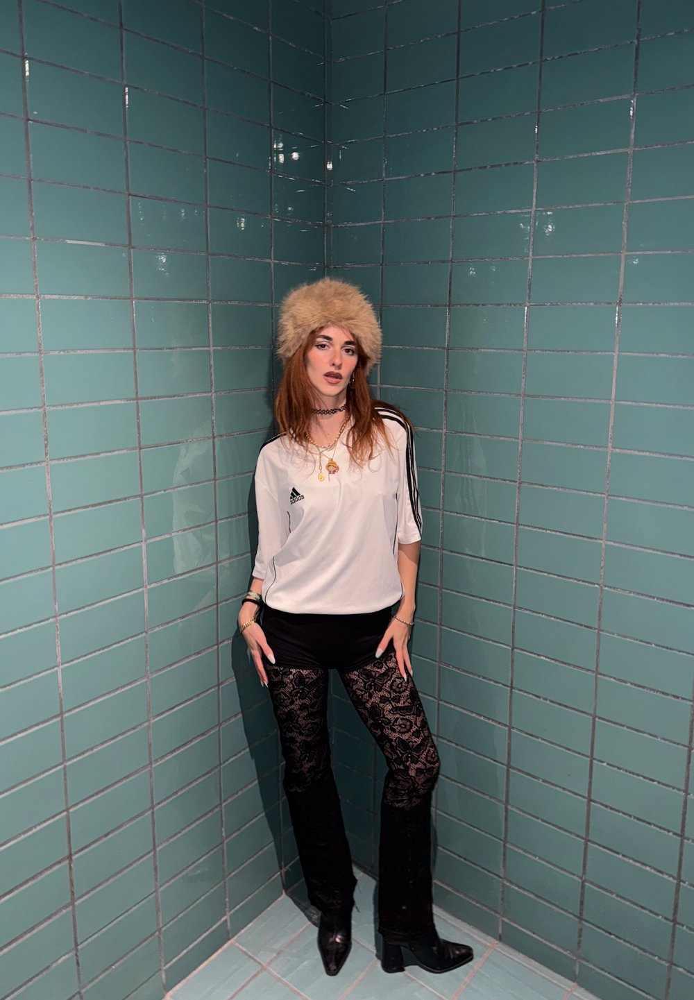 Valge spordisärk mustade triipudega, mustad lühikesed püksid ning pitsimustriga mustad püksid. Tan-nahavärvi karvamüts ja mustad kontsaga saapad. Roheline plaaditud taust.