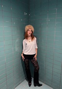 Valge spordisärk mustade triipudega, mustad lühikesed püksid ning pitsimustriga mustad püksid. Tan-nahavärvi karvamüts ja mustad kontsaga saapad. Roheline plaaditud taust.