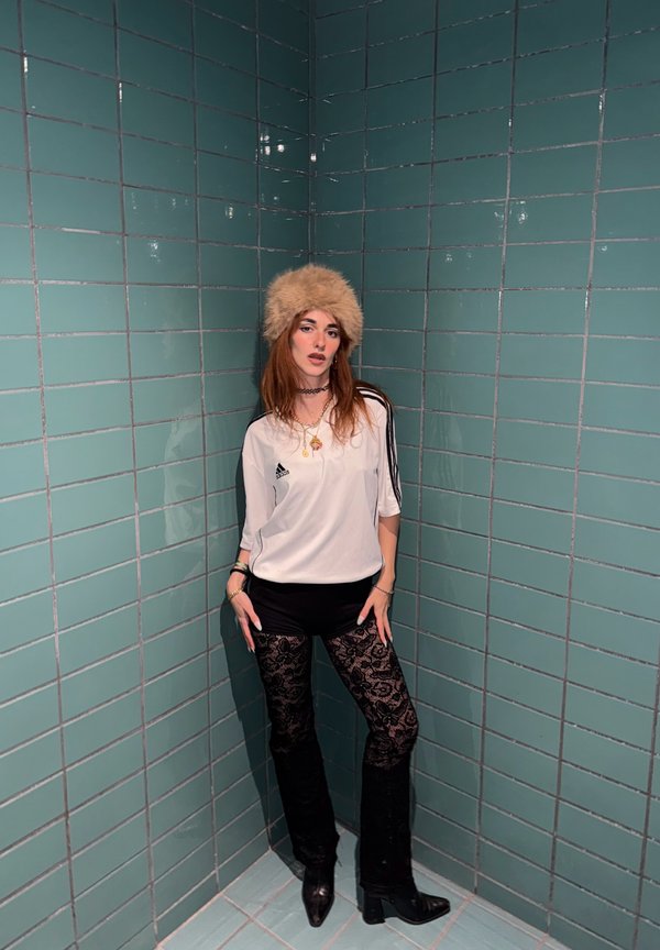 White sports shirt with black stripes, black shorts, and black lace-patterned trousers. Tan fur hat and black heeled boots. Green tiled background.