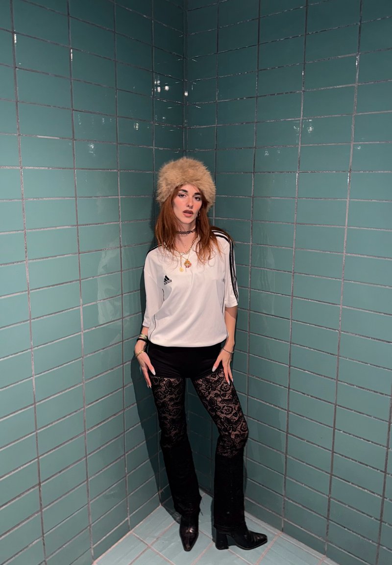 Chemise de sport blanche à rayures noires, short noir et pantalons noirs à motif de dentelle. Chapeau en fourrure beige et bottes noires à talons. Fond en carreaux verts.