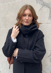 Vrouw met schouderlang rood haar, gekleed in een zwarte jas met grote knopen, terwijl ze de kraag vasthoudt, staand tegen een lichte stenen muur.