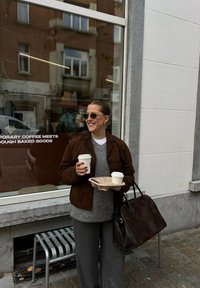 Καφέ σουέντ μπουφάν, γκρι πουλόβερ και φαρδιά γκρι παντελόνια· κρατώντας δύο κούπες καφέ σε μια χαρτοσακούλα και μια σκούρα τσάντα. Αστική τοποθεσία.