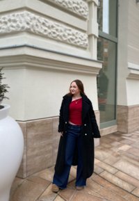 Μαύρο χνουδωτό παλτό πάνω από κόκκινο τοπ, συνδυασμένο με τζιν φαρδιά παντελόνια και παπούτσια με τακούνια. Στηρίζεται σε πλακόστρωτη επιφάνεια με μια λευκή γλάστρα κοντά.