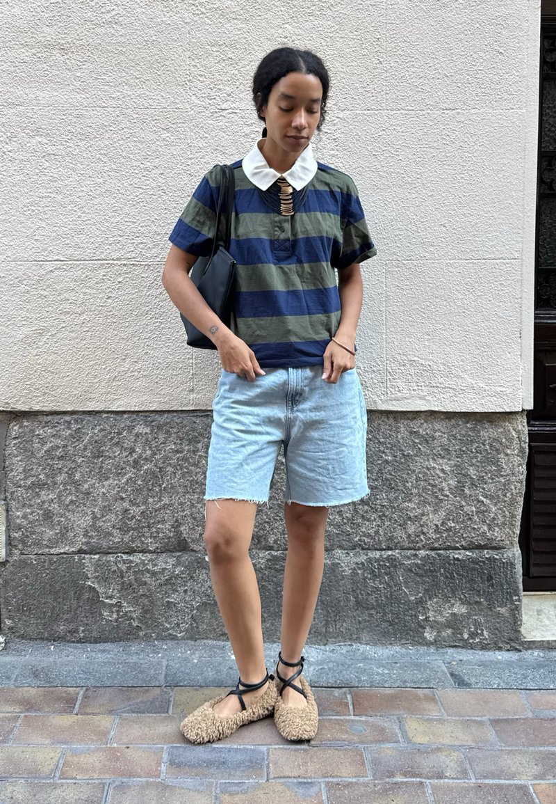 Donna in piedi all'aperto, vestita con una maglietta a righe con colletto bianco, pantaloni corti in denim chiaro, scarpe beige pelose e che porta una borsa a tracolla nera.