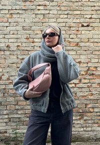 Casaco bomber cinza com fecho em zip e punhos canelados, cachecol cinza em camadas, blusa preta, jeans escuros e mala rosa em trama. Fundo de tijolo.