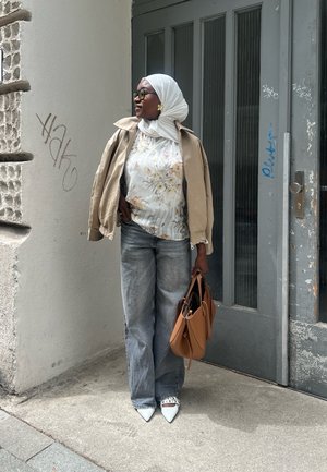 Mujer vestida con una chaqueta beige sobre una blusa floral, jeans grises de pierna ancha, zapatos blancos de punta con tachuelas y sosteniendo un bolso tote marrón.