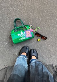 Personne portant des chaussures noires à orteils fendus et un jean effiloché se tient sur le trottoir près d'un sac Adidas vert avec des porte-clés, des lunettes de soleil noires et une bague jaune.