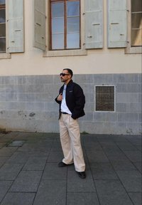 Schwarze Jacke mit Reißverschluss, weißes Hemd und beigefarbene, weit geschnittene Hosen. Sonnenbrille und schwarze Schuhe. Hintergrund mit strukturiertem Steinmauer.