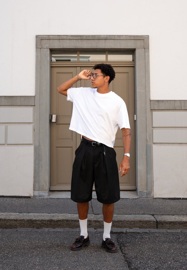 Jeune homme portant des lunettes, t-shirt blanc, short noir à fines rayures, chaussettes blanches et chaussures foncées, debout en ajustant ses lunettes devant une porte beige.