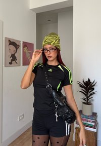 Joven mujer que lleva puesta ropa deportiva negra de Adidas, gorro verde con estampado, gafas y bolsa de hombro negra en una habitación con arte y libros.