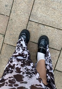Masywne czarne loafersy z srebrnym akcentem, zestawione z przezroczystą, brązowo-białą spódnicą w kropki, leżące na kamiennej nawierzchni.