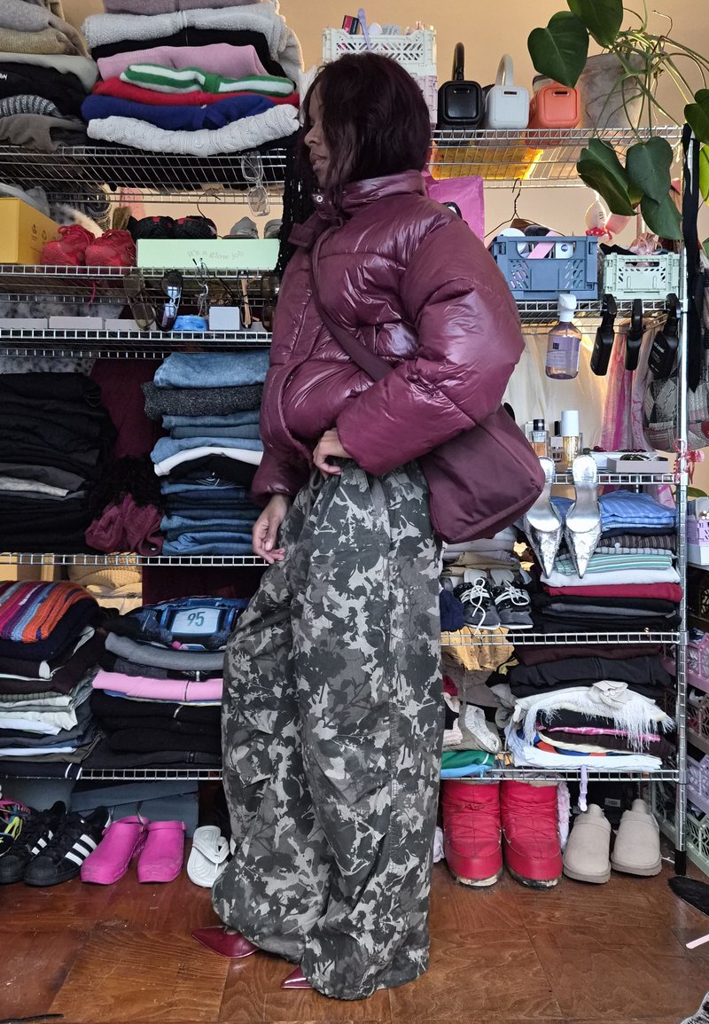 Puffy maroon jacket with wide sleeves paired with oversized camouflage pants. Various folded clothing stacked on metal shelves in the background.