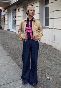 Lichtbeige leren bomberjack, zwart-roze gestreept poloshirt, wijd uitlopende donkere denim jeans en bruine schoenen. Stedelijke achtergrond.