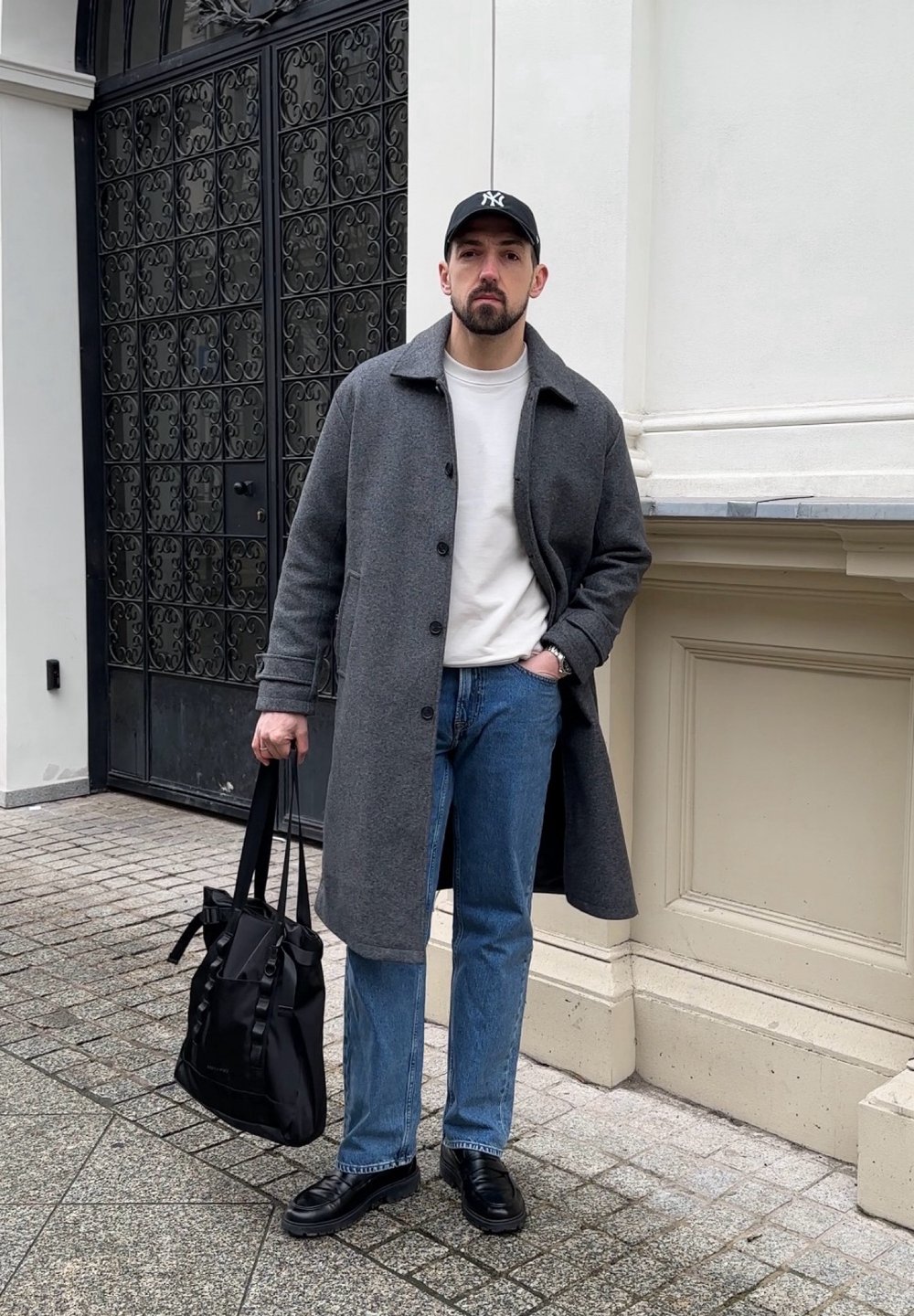 Cappotto di lana grigio sopra una camicia bianca, jeans blu, scarpe nere e con una borsa nera in mano. Lo sfondo mostra cancelli in metallo nero ornati.