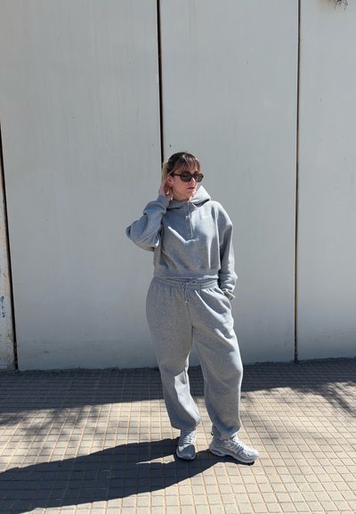 Mujer con sudadera con capucha y pantalones deportivos grises, con gafas de sol, está de pie en una acera adoquinada contra una pared clara, una mano tocando el cuello, la otra en el bolsillo.