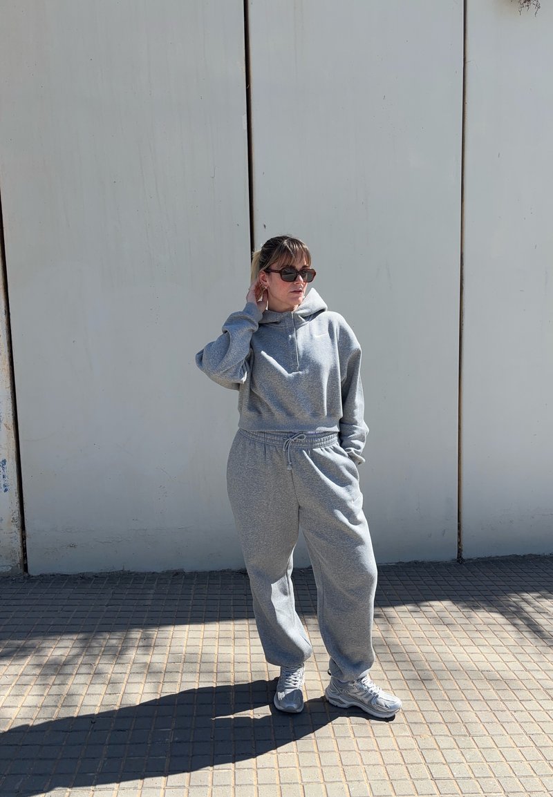 Γυναίκα με γκρι φούτερ με κουκούλα και φόρμα, φορώντας γυαλιά ηλίου, στέκεται σε πλακόστρωτο πεζοδρόμιο μπροστά σε φωτεινό τοίχο, ένα χέρι αγγίζει τον λαιμό, το άλλο στην τσέπη.