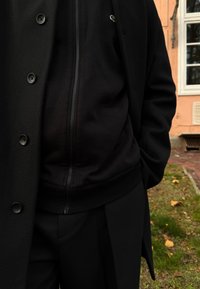 Črni, teksturiran plašč z gumbi čez črno jopico z zadrgo in črnim hlačam. Material je videti mehak in ustvarja večplastni videz.