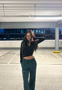 Schwarzes, geripptes Crop-Top mit Reißverschluss, grüne, locker sitzende Hose mit elastischem Bund und runde Brille; steht in einer Parkgarage.