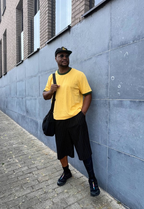 Homme portant un t-shirt jaune, un short à fines rayures, une casquette noire et des lunettes de soleil, debout contre un mur gris carrelé sur un trottoir pavé.