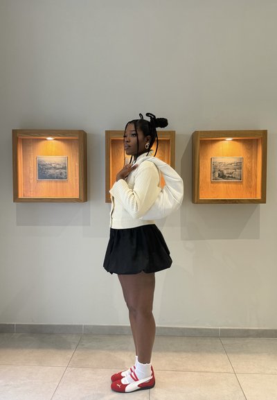 Joven mujer con jersey blanco, falda negra y zapatillas rojas está de perfil sosteniendo una bolsa blanca en una galería con fotos enmarcadas en la pared.