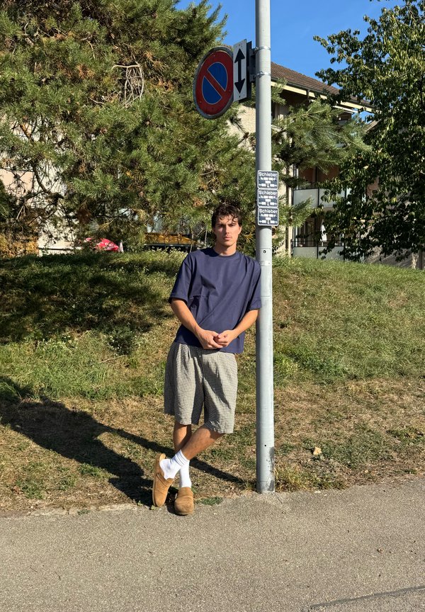 Jeune homme en chemise bleu et short à carreaux appuyé contre un poteau avec un panneau d'interdiction de stationner par une journée ensoleillée près de l'herbe et des arbres.