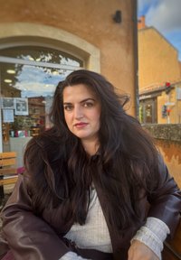 En kvinde med langt sort hår bærer en mørkebrun læderjakke over en lys grå sweater. Hun sidder foran et butiksvindue.