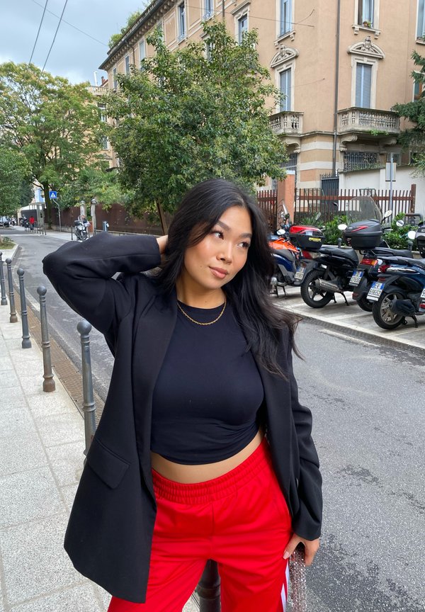 Blazer noir sur un crop top noir ajusté et un pantalon rouge à taille élastique. Cheveux texturés, collier en chaîne dorée, arrière-plan urbain.