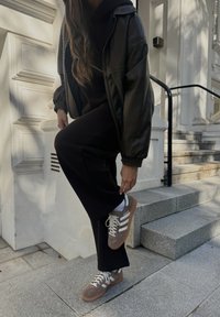 Μαύρη βομβιστική τζάκετ πάνω από μια σκούρα φούτερ, συνδυασμένη με μαύρο παντελόνι και καφέ αθλητικά παπούτσια με λευκές ρίγες και μπεζ λεπτομέρειες, που στέκεται σε πέτρινες σκάλες.