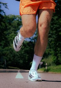 Ténis de desporto brancos com riscas pretas, usados com meias brancas, combinados com calções laranja. A imagem mostra apenas as pernas em movimento num caminho.
