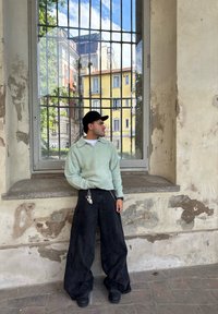 Mintgrön fluffig tröja, breda svarta jeans, svart keps, står vid ett fönster med utsikt över byggnader. Notera den avskalade väggtexturen.