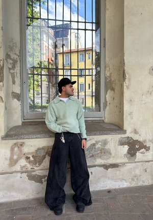 Jersey de lana verde menta, pantalones de mezclilla anchos negros, gorra negra, de pie cerca de una ventana con vista a los edificios. Nota la textura de las paredes desconchadas.