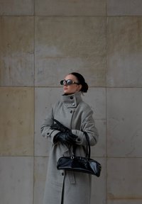 Šedý vlněný kabát s vysokým límcem, černé kožené rukavice a černá kabelka. Doplňky zahrnují velké tmavé sluneční brýle. Texturované kamenné pozadí.