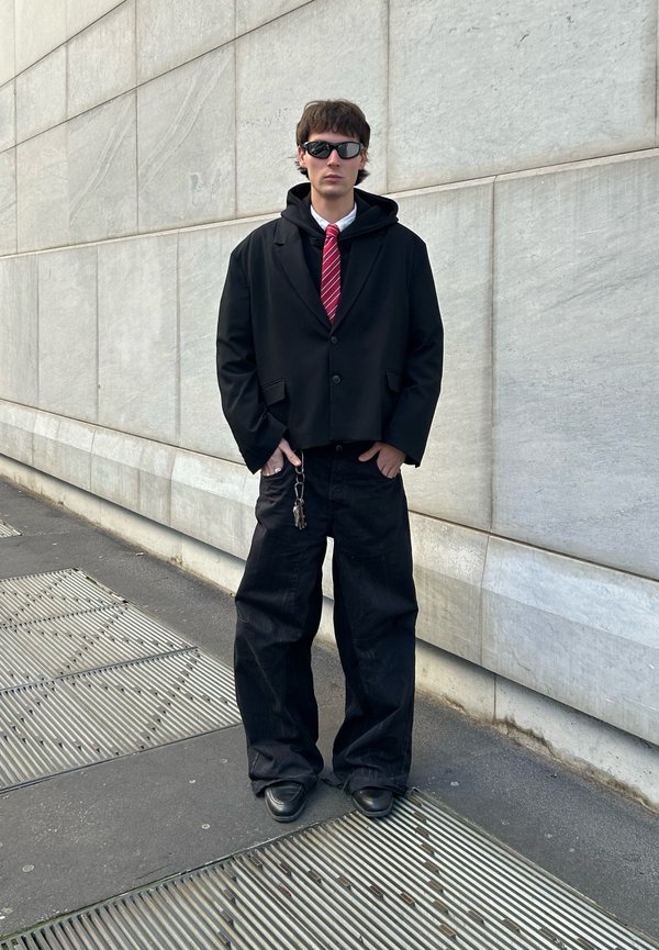 Zwarte oversized blazer over een grijze hoodie, gecombineerd met zwarte baggy broeken en zwarte schoenen. Rood-witte gestreepte stropdas. Donkere zonnebril.