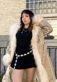 Beige nepbontjas met witte voering; zwarte top en uitlopende zwarte rok; zilveren hanger riem; textuur handtas. Kenmerken zijn lange mouwen en pluizige afwerking.