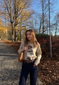 Béžový sveter s pleteným vzorom medveďa, tmavomodré široké nohavice, hnedý opasok a hnedá kabelka, na pozadí cesty v jesennej krajine.