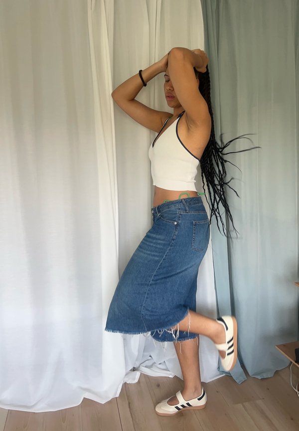 Bela rebrasta kratka majica z mornarsko modrim robom, modra denim midi krila z raztrgano obrobo in smetanaste čevlje z črnimi črtami, ki stoji ob draperijah.