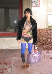 Jeune femme aux cheveux noirs courts portant une veste en fausse fourrure noire, un haut à motifs floraux, des collants bleus, des chaussures à motif léopard, tenant un sac Hello Kitty violet en extérieur.