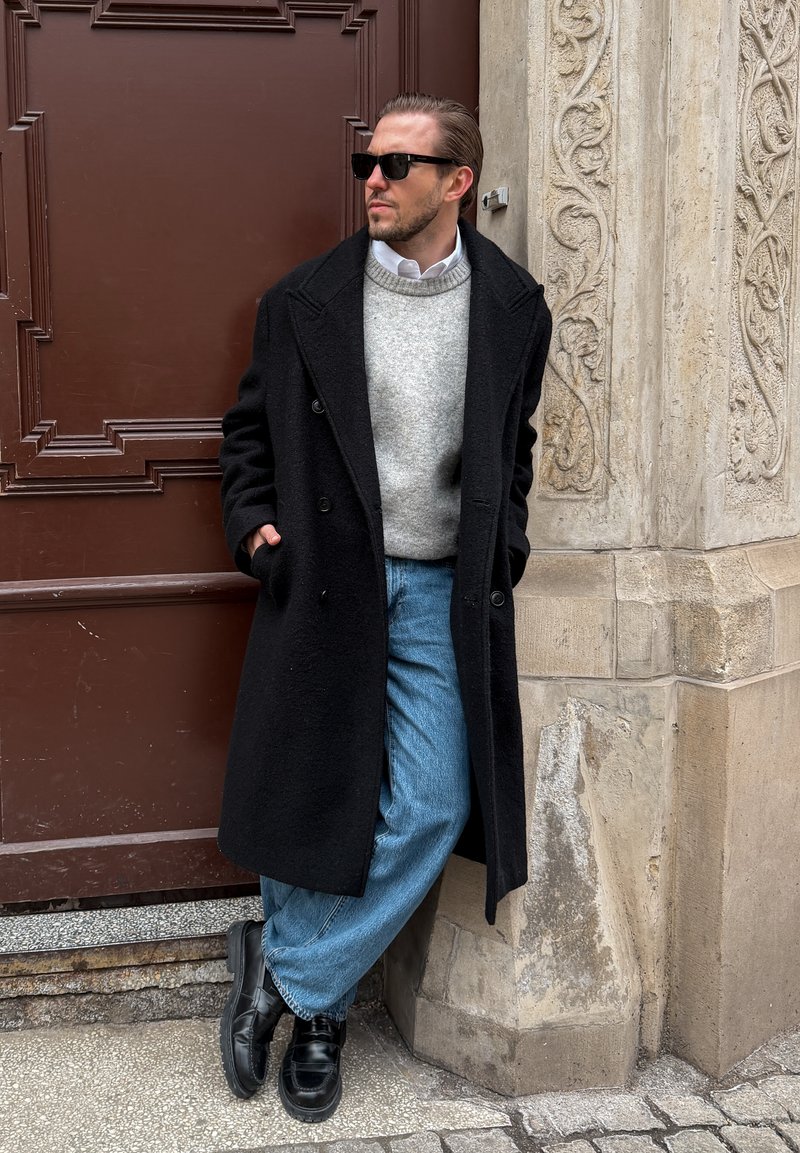 Homme portant un pardessus noir, un pull gris, un jean bleu et des lunettes de soleil, appuyé contre un mur en pierre ornée près d'une porte marron.